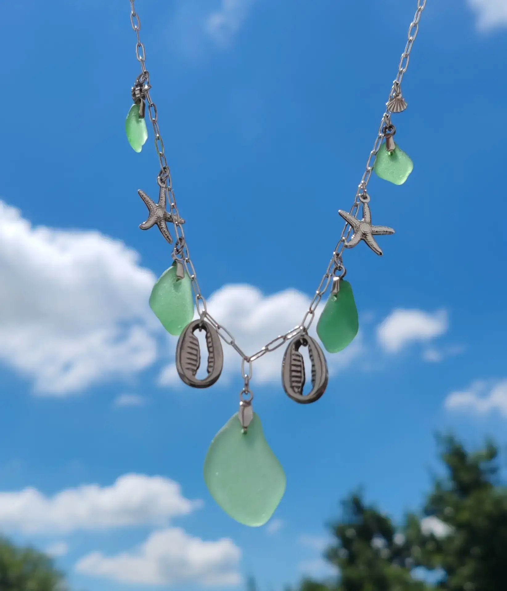 Le collier Rivage tenu devant un ciel bleu, ses éclats de verre de mer verts et ses cauris en pleine lumière