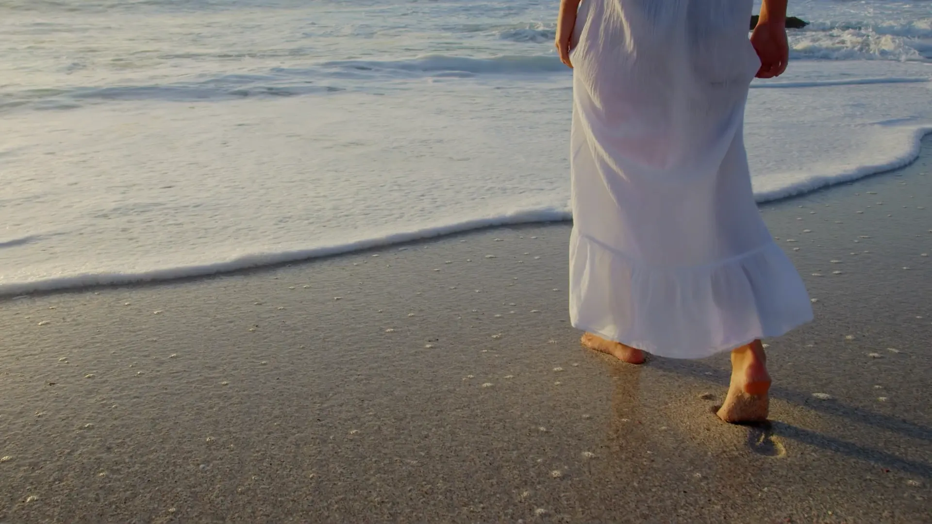 Une femme en robe blanche marche pieds nus au bord de l’eau, à l’heure où la mer se retire sur le sable